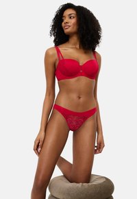 Görbe hajú nő piros csipke fehérneműben, egyik térdét egy kerek bézs párnán támasztva pózol egy sima háttér előtt.