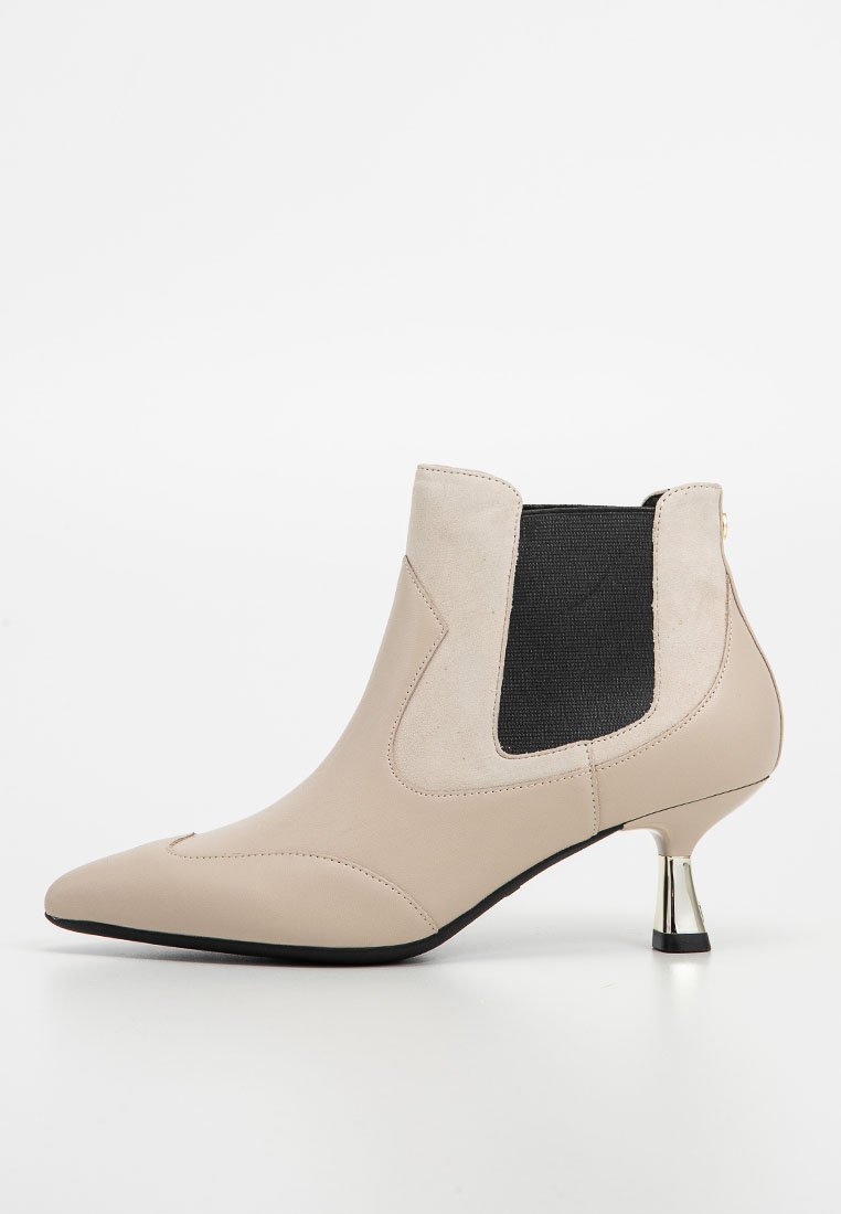 Botín beige hasta el tobillo con punta afilada, paneles laterales elásticos y un tacón en forma de cono de color plateado brillante. Presenta cuero suave y acentos texturizados.