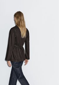 Žena s dlouhými blond vlasy, oblečená v tmavě hnědém sako se páskem a tmavě modrých džínách, kráčející proti jednoduchému světlému pozadí, otočená zády.