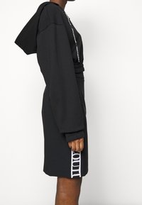 Osoba v černé mikině s kapucí a černé sukni s bílým vertikálním textem na boku, viděná z boku na jednoduchém pozadí.