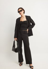 Melna formāla apģērba komplekts, kas sastāv no pieguļošas blūzes, piegriezta žaketes un platām biksēm. Aksesuāri ietver melnus saulesbrilles un elegantu rokas somu.