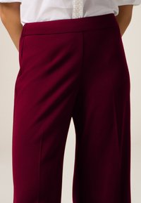 Pantaloni tailored color bordeaux con dettagli di pieghe nette, realizzati in un tessuto liscio; vita alta con una finitura discreta e taglio dritto.