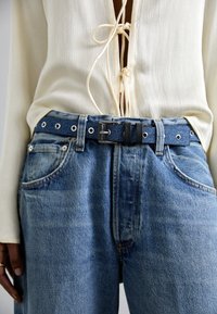 Blusa de color claro y texturizada con escote en V profundo y detalle de lazo, combinada con jeans de denim azul con costuras visibles y un cinturón de denim.