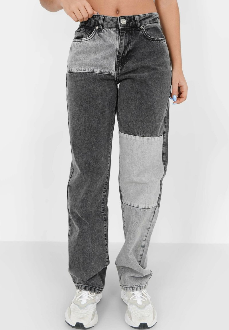 Personne portant un jean patchwork gris bicolore et des baskets blanches, debout devant un fond clair uni.