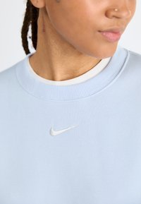 Světle modrá mikina vyrobená z měkkého materiálu s žebrovaným výstřihem a malým bílým logem Nike na hrudi. Čistý design a minimální detaily.