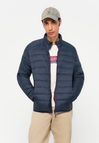 Marineblå dunjakke med quiltet design, høj krave og kontrastfarvet rød lynlås. Båret over en lyse t-shirt og beige bukser. Kasket tilføjer et afslappet detaljer.