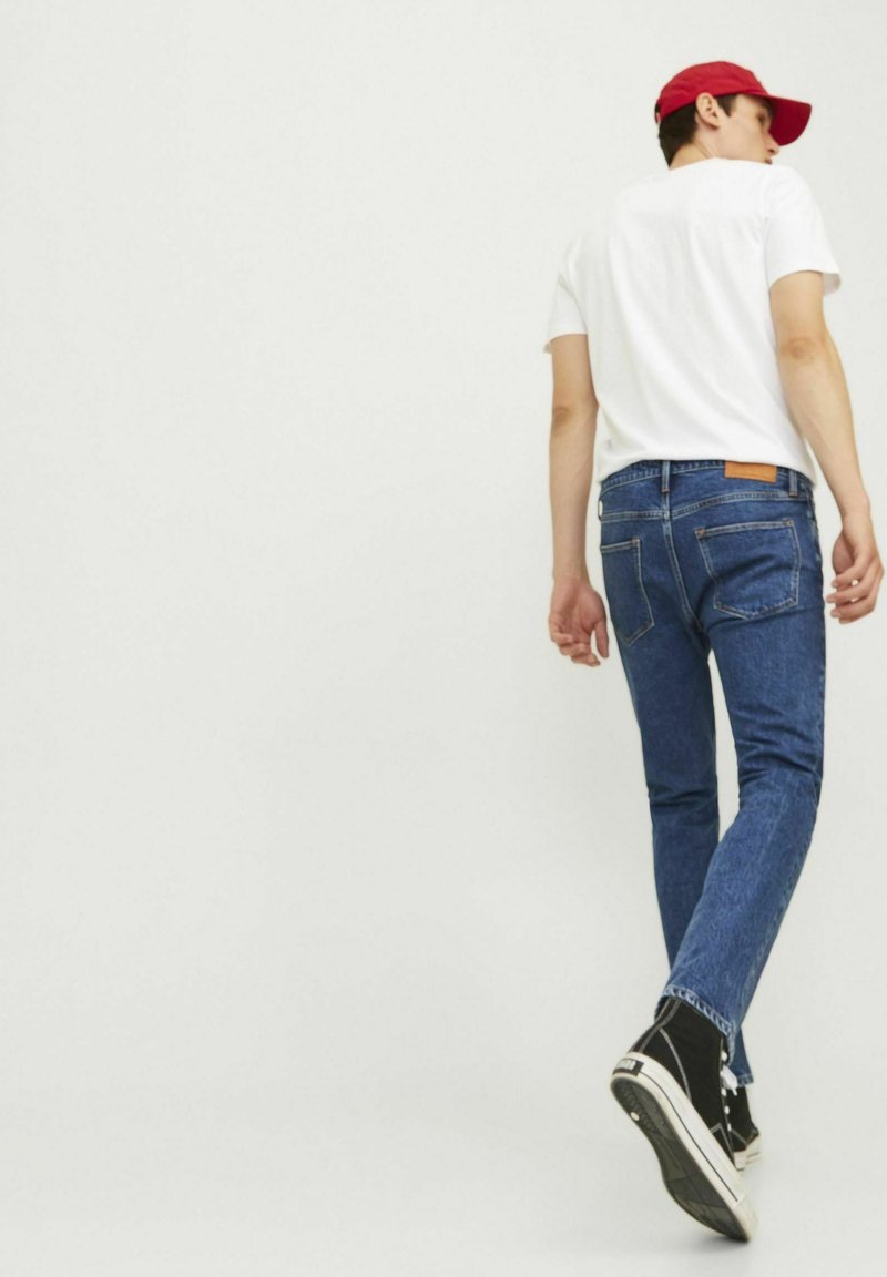 Jeans de mezclilla azul con un ajuste slim, con bolsillos traseros, un parche de cuero marrón y combinados con una camiseta blanca y zapatillas altas negras.