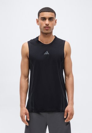 Młody mężczyzna z krótkimi włosami, ubrany w czarną sportową koszulkę bez rękawów marki Adidas oraz szare spodenki, stojący na jednolitym białym tle.