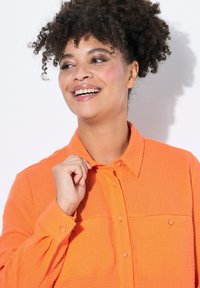 Camicia a bottone arancione con texture, colletto e due tasche sul petto. Modello che sistema leggermente il colletto, sorridendo su uno sfondo bianco.