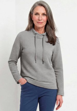Vrouw met grijs haar draagt een gestructureerde grijze longsleeve met trekkoorden en blauwe broek, staande met één hand in de zak.