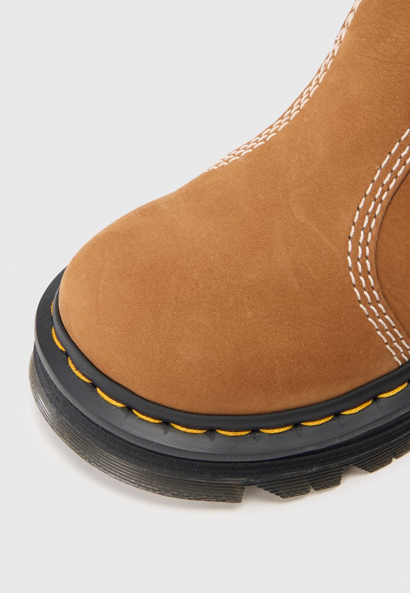 Brauner Wildlederstiefel mit kontrastierendem weißen Stich, runder Spitze, schwarzer Gummisohle und gelben Akzenten am Rand. Texturierte Oberfläche.