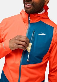 Veste de course colorée en matériau imperméable, avec une partie supérieure orange et une partie inférieure bleue, poche zippée et capuche ajustable.