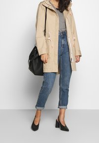 Beżowa parka z kapturem na sznurek, zestawiona z szarą bluzką i dżinsami z wysokim stanem. Całość dopełnia czarna torebka i czarne buty na obcasie.