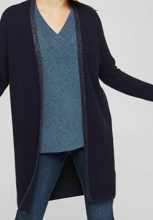 Vrouw die een marineblauwe open cardigan met ribstructuur draagt over een blauwe V-hals trui en blauwe spijkerbroek, met licht gespreide armen.