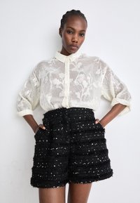 Blusa blanca transparente con patrón floral y cierre de botones, combinada con pantalones cortos negros texturizados con rayas horizontales y acentos plateados.