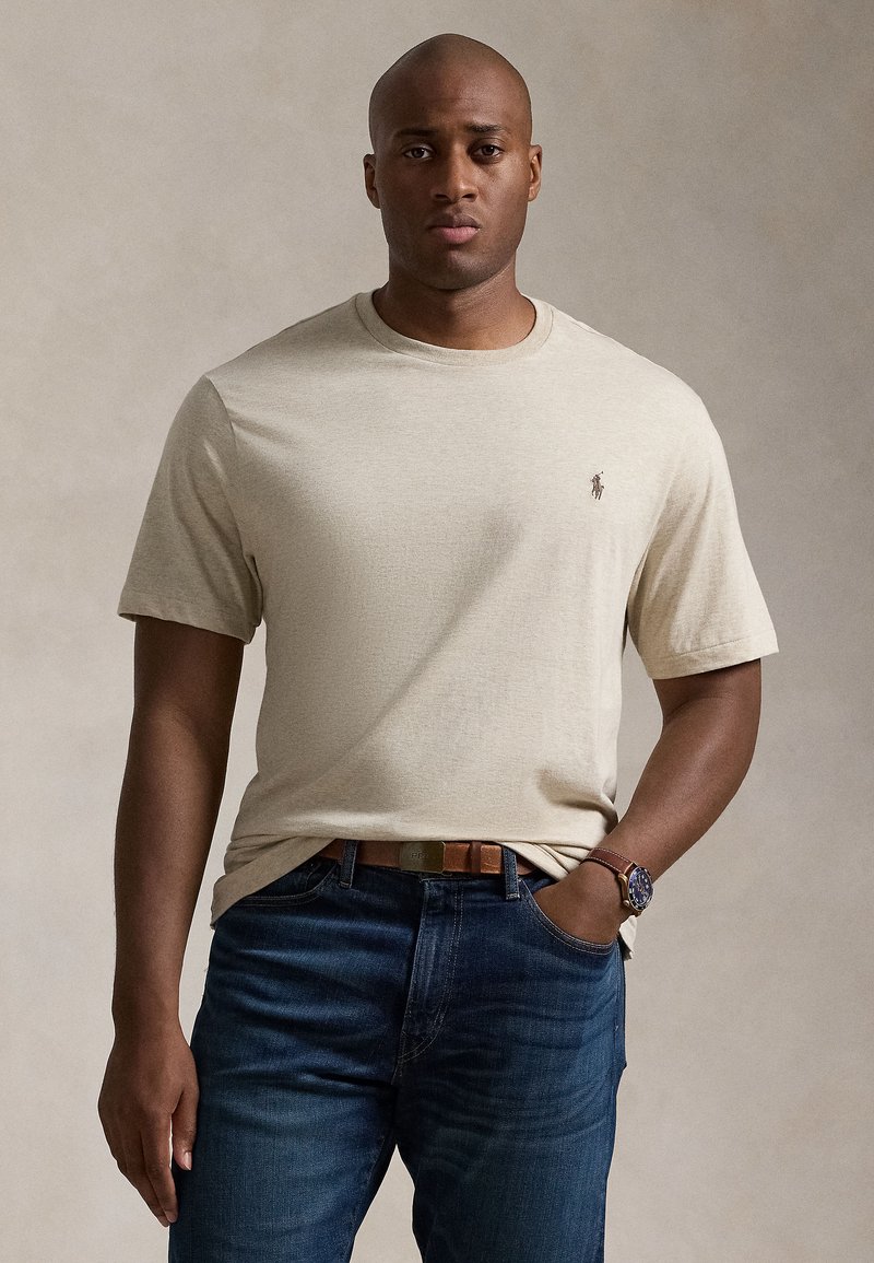 Beige t-shirt med korta ärmar och rund hals, med en liten logotyp på vänster bröst. Kombinerad med blå jeans och ett brunt skärp.