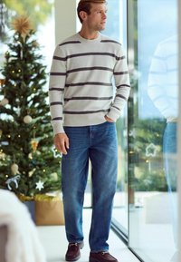 Gray and cream striped sweater, blue denim jeans, and dark brown shoes. Standing near a window with a decorated Christmas tree in the background.