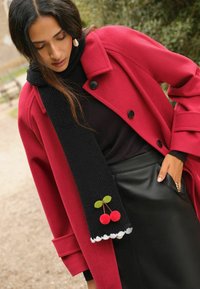 Červený vlněný kabát, černý žebrovaný šátek s aplikací třešně, černá sukně z umělé kůže a strukturované detaily, stylizováno pro vrstvený vzhled.