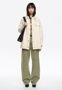 Chaqueta de camisa de lana crema con botones de presión, dos bolsillos en el pecho, combinada con pantalones de pana verde oliva de pierna ancha y botas negras de tacón.