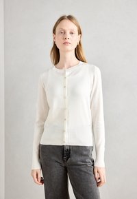 Bijela pletena cardigan jakna s okruglim izrezom, zatvaranjem na gumbe sprijeda i dugim rukavima. Ima rebrasti rub i mekan, lagan tekstil.