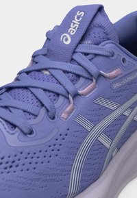 Κοντινό πλάνο πορφυρού παπουτσιού τρεξίματος Asics Gel-Cumulus με λεπτομερείς κορδόνια, πλέγμα στο πάνω μέρος και λογότυπο της εταιρίας στη γλώσσα και στην πλευρά.
