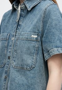 Πουκάμισο από τζιν σε ανοιχτό γαλάζιο με κοντά μανίκια, δύο τσέπες στο στήθος, ασημένια μεταλλικά στοιχεία και λογότυπο στην τσέπη. Υφασμάτινη υφή.