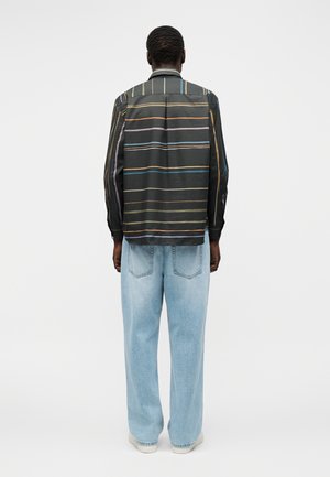 Personne portant une chemise noire à manches longues avec des rayures horizontales multicolores et un jean bleu clair ample, debout de dos.