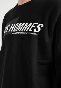 Sudadera negra con cuello redondo, que presenta un texto blanco en negrita: "LES HOMMES" y un texto más pequeño debajo. Tejido suave con textura mínima.