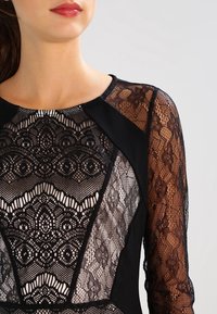 Černé šaty s propracovanými krajkovými detaily a dlouhými černými krajkovými rukávy, s pevným černým tělem a přiléhavým střihem.