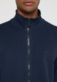 Chaqueta navy con cremallera y cuello alto, de tejido texturizado, y un pequeño logo en el pecho. Cuenta con una cremallera de longitud completa con dientes metálicos.