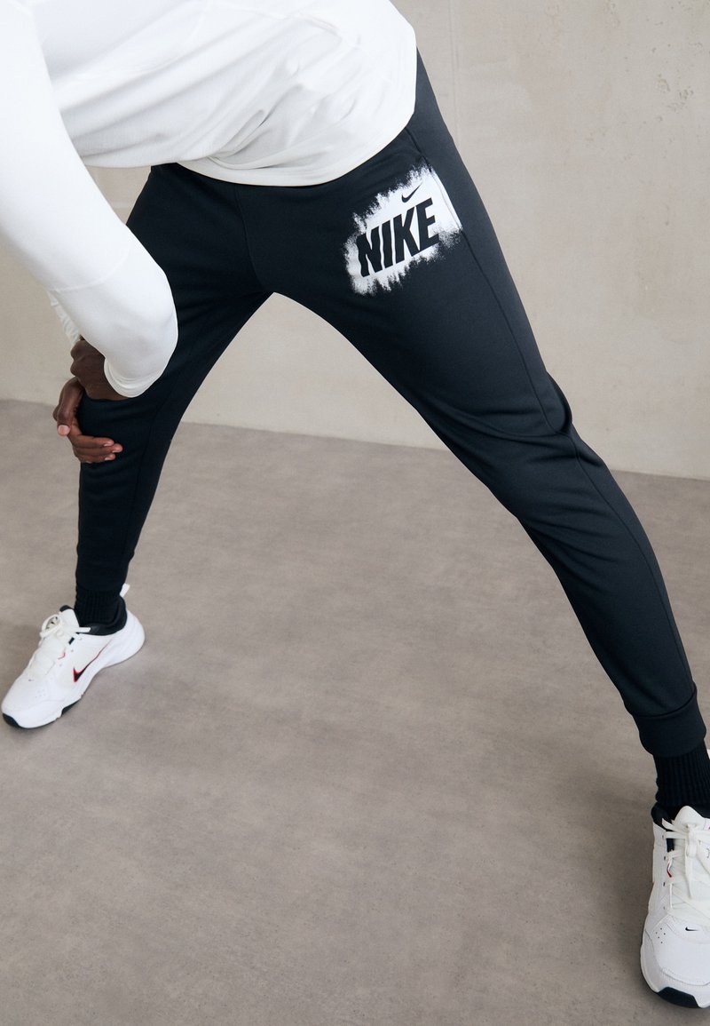 Nike mustat joggerit, joissa on joustava vyötärö, kapeneva muotoilu ja valkoinen logo reidessä, yhdistettynä valkoisiin urheilukenkiin.