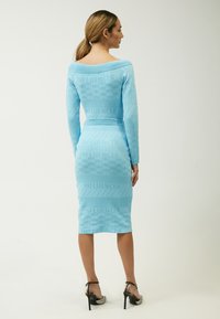 Abito azzurro chiaro con maniche lunghe, tessuto testurizzato, silhouette aderente, caratterizzato da un motivo ripetuto "INFLUENCER". Abbinato a sandali neri con tacco.