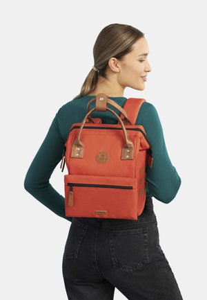Zaino in tela arancione con dettagli in pelle marrone, forma rettangolare, tasca con zip frontale, patch del logo e manici superiori con una tracolla.