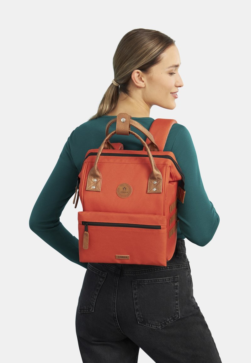 Zaino in tela arancione con dettagli in pelle marrone, forma rettangolare, tasca con zip frontale, patch del logo e manici superiori con una tracolla.