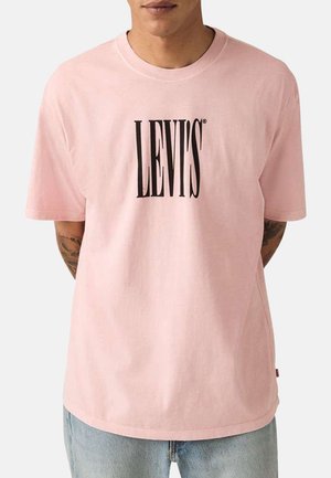Hombre vistiendo una camiseta Levi's de color rosa claro con texto de logo en negro negrita, combinada con pantalones vaqueros azul claro sobre un fondo liso.
