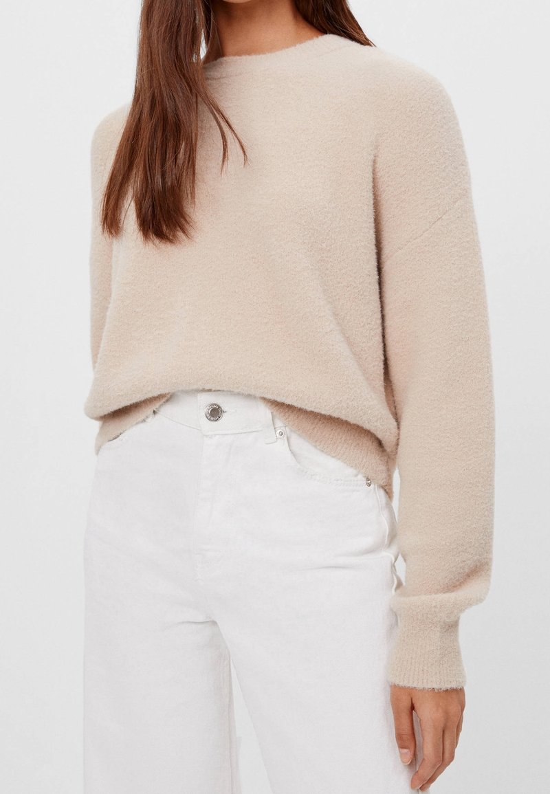 Femme portant un pull en tricot beige doux et un jean blanc taille haute, debout avec la main posée de façon détendue à côté du corps.