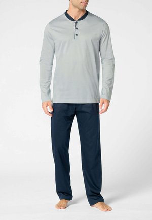 Uomo che indossa una camicia henley a maniche lunghe grigio chiaro con rifiniture blu scuro e pantaloni larghi blu scuro, in piedi a piedi nudi su uno sfondo bianco.