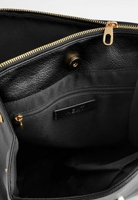 Intérieur d'un sac à main en cuir noir avec une poche zippée dorée et un fermoir à pression, montrant un revêtement texturé et une petite étiquette en cuir de marque.