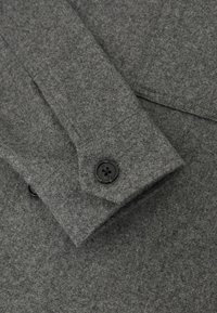 Grijze viltstof met een gladde textuur, voorzien van een zwarte knoopsluiting. Detail bevat een omgevelde manchet.