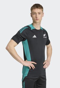 Camisa deportiva negra con acentos turquesa, con mangas cortas, cuello redondo y tres detalles a rayas en cada hombro. Logotipo de Adidas visible.
