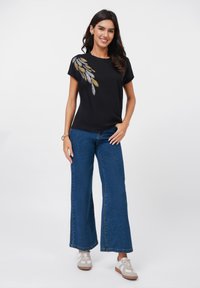 T-shirt noir à manches courtes avec broderie en feuilles dorées et argentées ; associé à un jean en denim bleu à jambes larges ; des baskets beiges complètent la tenue.