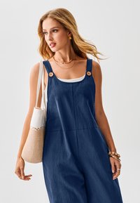 Marineblauer Denim-Jumpsuit mit weitem Bein, quadratischem Ausschnitt, Knopfdetails, getragen mit weißem Oberteil und Häkel-Tasche. Natürliche Haare.