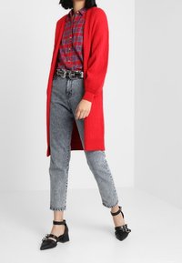 Rode geribbelde cardigan over een geruite blouse, gecombineerd met grijze denim jeans met hoge taille en zwarte schoenen met blokhak en banden.