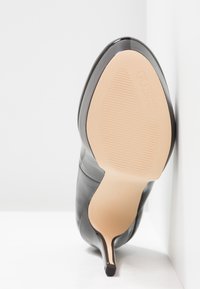 Chaussure noire à talon haut avec une finition brillante, bout pointu et semelle en caoutchouc beige texturée. Contient un logo sur la semelle.