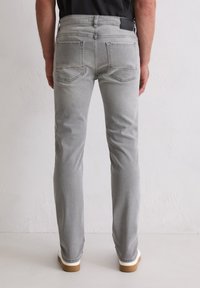 Homme portant un jean gris slim-fit et des baskets blanches, se tenant contre un mur blanc uni avec les mains détendues sur les côtés.