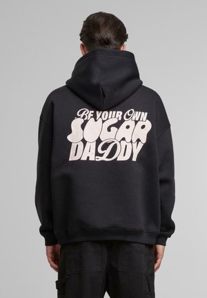Black hoodie made of cotton, featuring a large, rounded graphic text "Be Your Own Sugar Daddy" in cream color on the back.