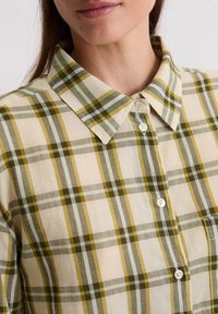 Collared plaid shirt in beige, green, and light blue. Cotton material with a button-down front and a lightweight texture.
