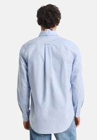 Cămașă lungă cu mâneci de culoare albastru deschis, cu manșete cu nasturi, pliere la spate și margine curbată. Material din bumbac cu o textură netedă și un design simplu.