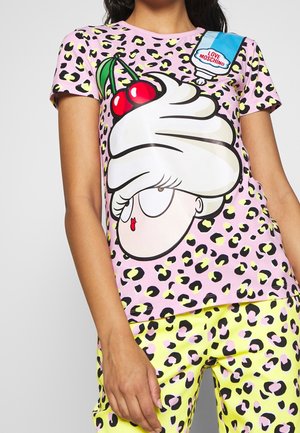 Mujer que lleva una camisa rosa con estampado de leopardo y un diseño de cara de caricatura con crema y cereza, combinada con pantalones amarillos con estampado de leopardo.