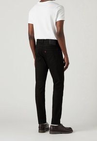 Jeans de denim negro con corte slim, que presentan bolsillos traseros y una pequeña etiqueta de logo roja. Combinados con una camiseta blanca y zapatos marrón oscuro.